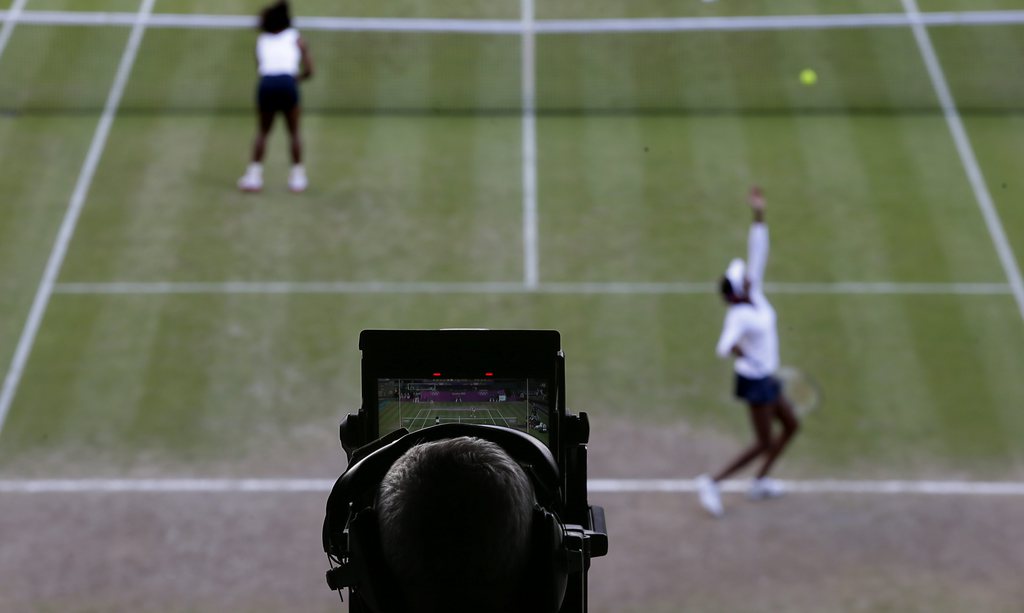 A TV cameraman focus on the court as Venus Williams, right, serves and Serena Williams, left, stands near the net during their women's doubles semifinal match against Maria Kirilenko and Nadia Petrova of Russia  at the All England Lawn Tennis Club at Wimbledon, in London, at the 2012 Summer Olympics, Saturday, Aug. 4, 2012. (AP Photo/Mark Humphrey)