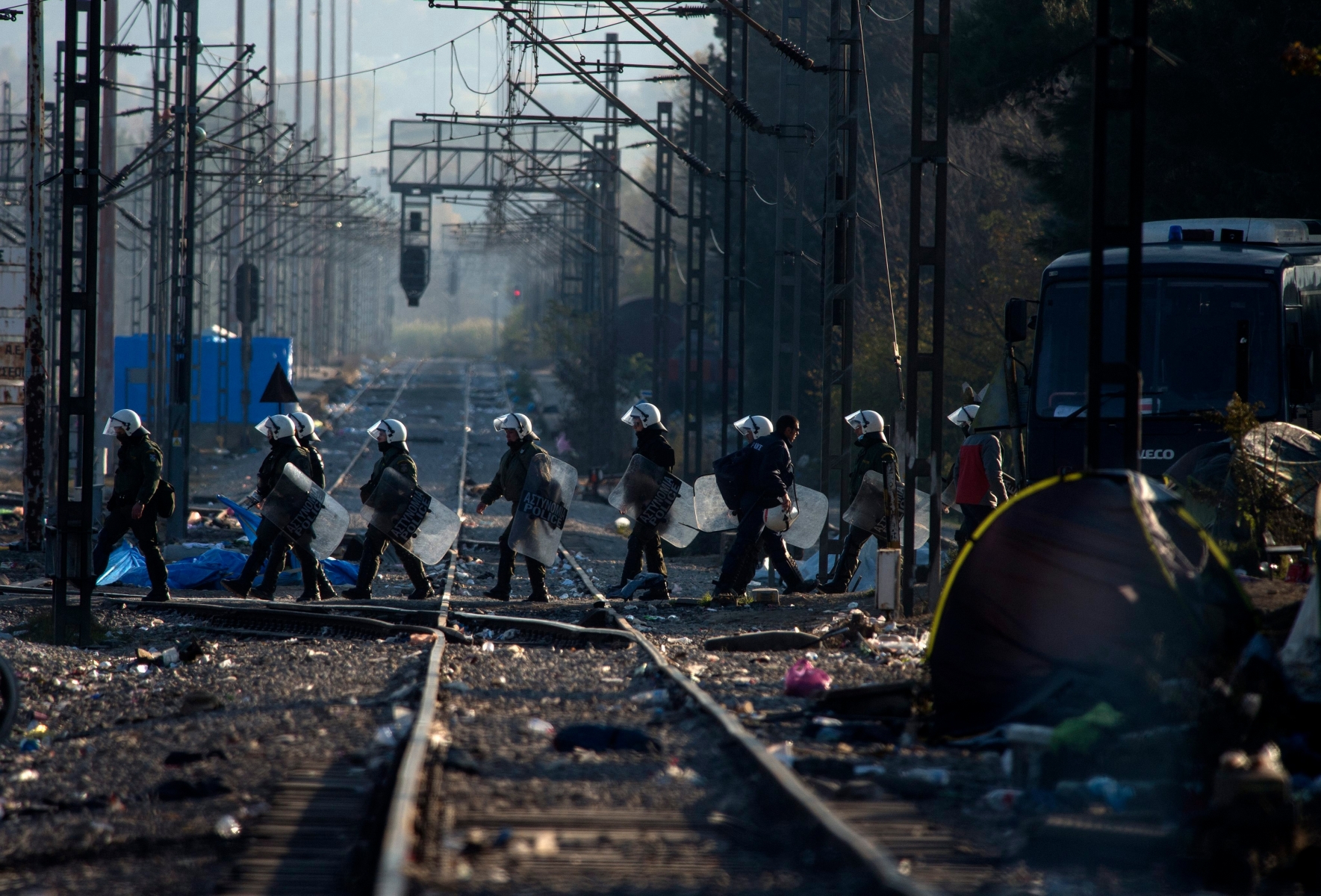 epa05061618 In a photo taken from the Macedonian side of the border, Greek police officers cross the rail tracks along the migrants camp near Idomeni, Greece, 09 December 2015. Greek police began dispersing crowds of people who have been stranded for weeks at Idomeni, a village on the country's border with Macedonia, local media reported. The migrants, mostly from Iran, Bangladesh, Pakistan and Morocco, were placed on buses, some forcibly, and transported to shelters in Athens 550 kilometres to the south, reports said. Around 1,200 people had been stranded at the border since Macedonia started denying entry to so-called economic migrants trying to reach Western Europe via the Balkan route, which stretches from Turkey, through Greece, Macedonia, Serbia, Croatia, Slovenia and finally to Austria. In Athens, they will have to apply for asylum or be repatriated. Greek police sealed off Idomeni on 09 December morning, also blocking access to reporters and humanitarian workers.  EPA/GEORGI LI