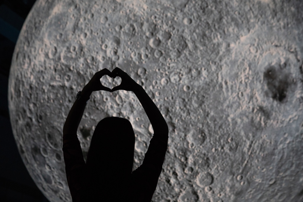 epa06954607 A Chinese woman makes a heart shape with her hands in front of a moon sculpture, the artwork by artist Luke Jerram, at the National Aquatics Center, known as the Water Cube, in Beijing, China, 17 August 2018. China marks Qixi Festival on 17 August, which is considered as Chinese Valentine's Day. Qixi Festival is celebrated on the seventh day of the seventh lunar month and is inspired by a Chinese folk tale of love between a cowherd and a fairy. EPA/ROMAN PILIPEY